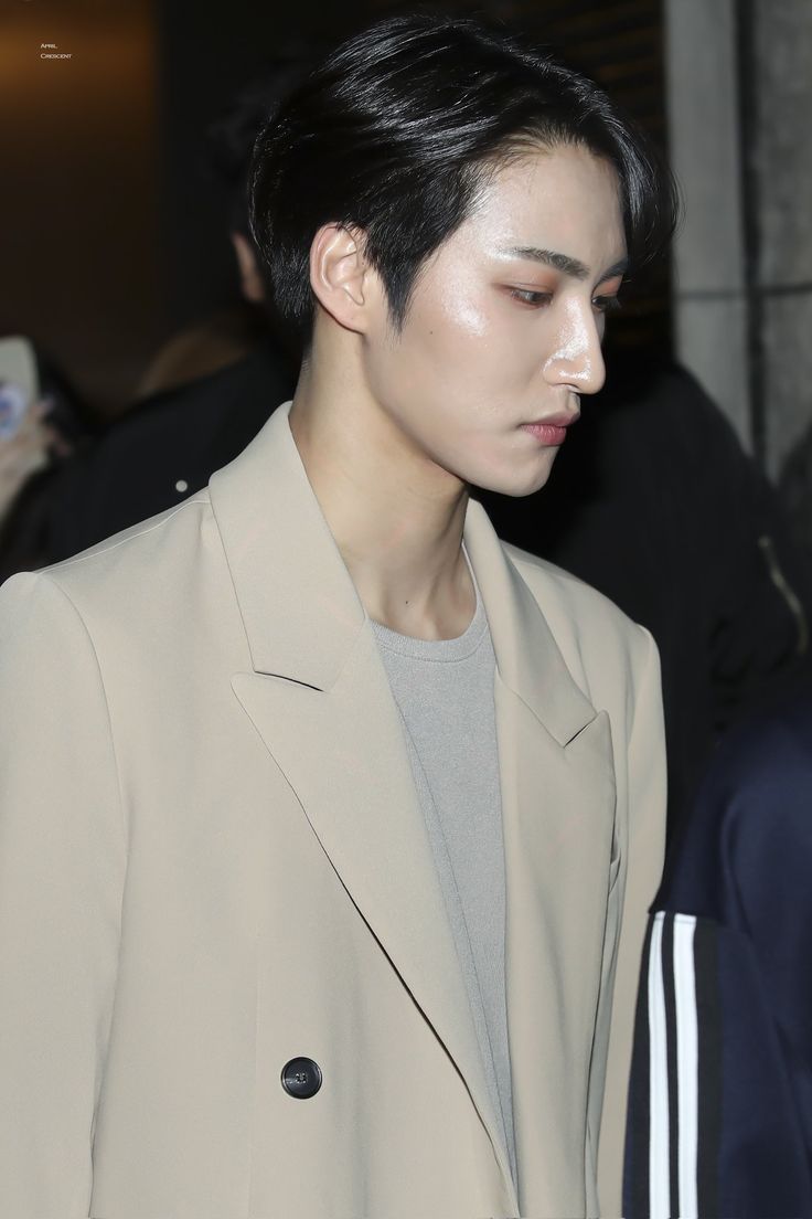 a man with black hair wearing a beige suit and grey shirt looking down at his cell phone