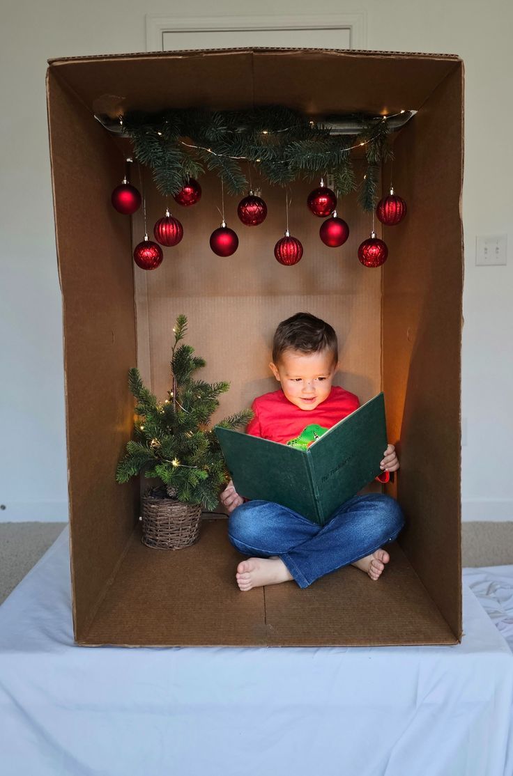 a young boy is sitting in a cardboard box reading a book with christmas decorations around him