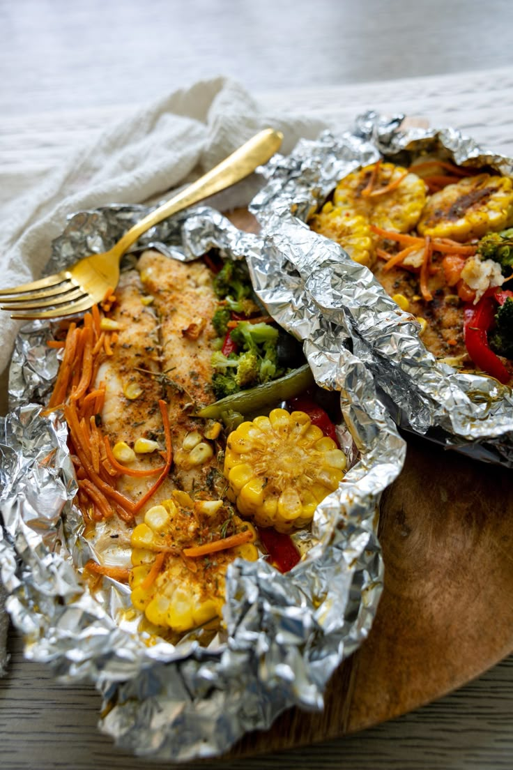 a wooden plate topped with chicken and vegetables covered in tin foil next to a fork