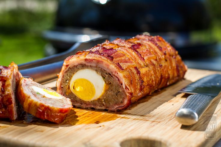 meatloaf wrapped in bacon and an egg on a cutting board next to a knife