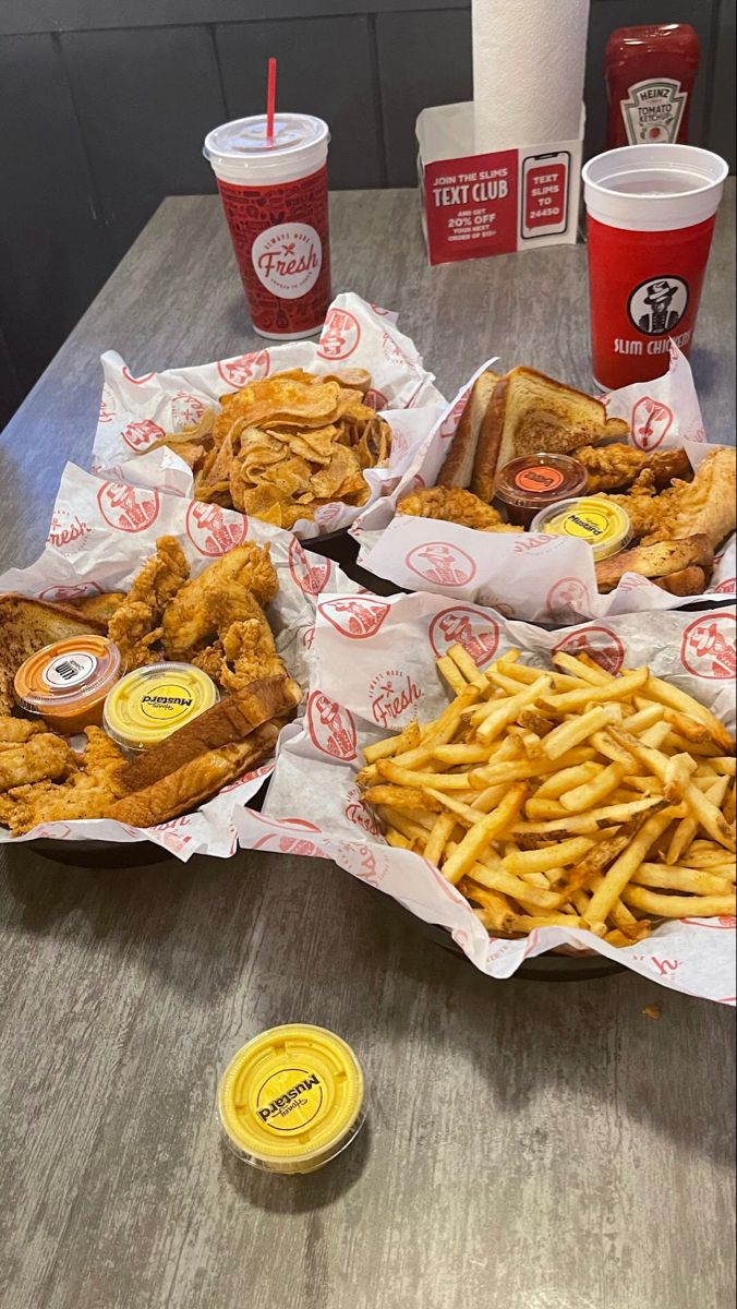 several trays of chicken and french fries on a table with two cups of soda