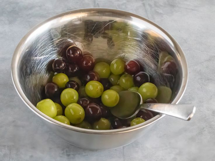 a metal bowl filled with olives on top of a marble countertop next to a silver spoon