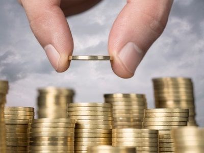 a hand is placing a coin on top of a pile of gold coins with clouds in the background