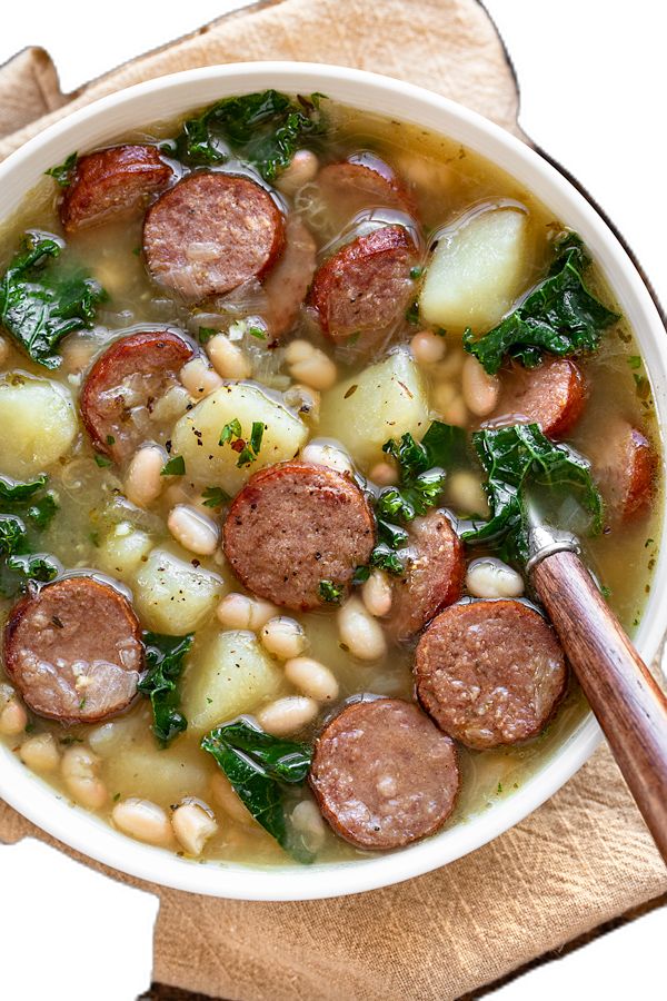 a white bowl filled with sausage, beans and greens next to a wooden spoon on top of a cutting board