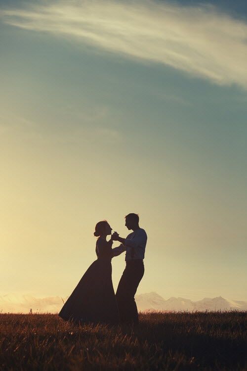 a man and woman standing in the grass with their arms around each other as the sun sets behind them