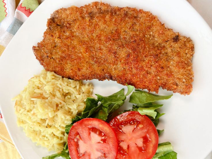 a white plate topped with meat and rice next to a slice of tomato on top of lettuce