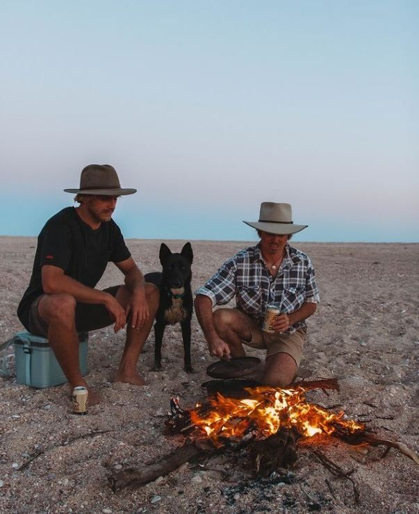 two men sitting around a campfire with a dog