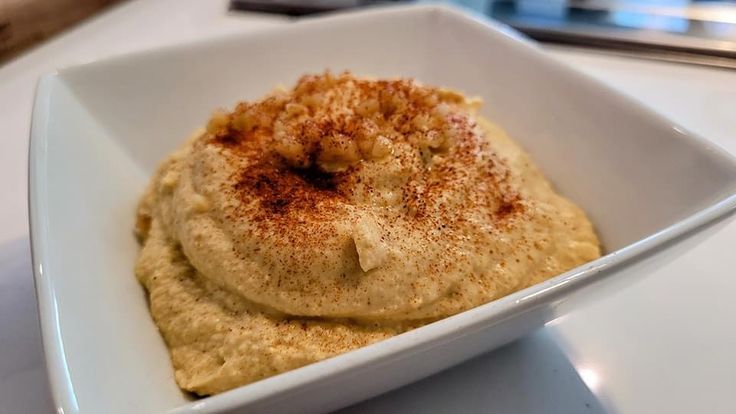 a square white bowl filled with hummus on top of a table next to a fork