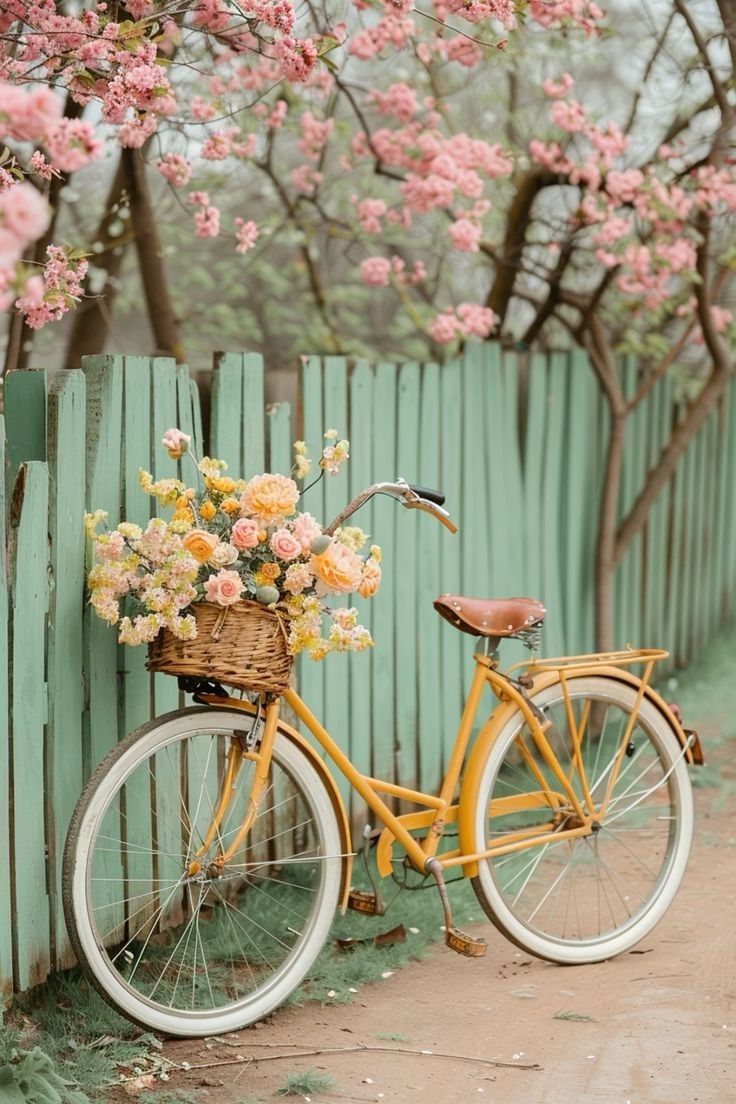 a yellow bicycle with flowers in the basket parked next to a green fence and tree