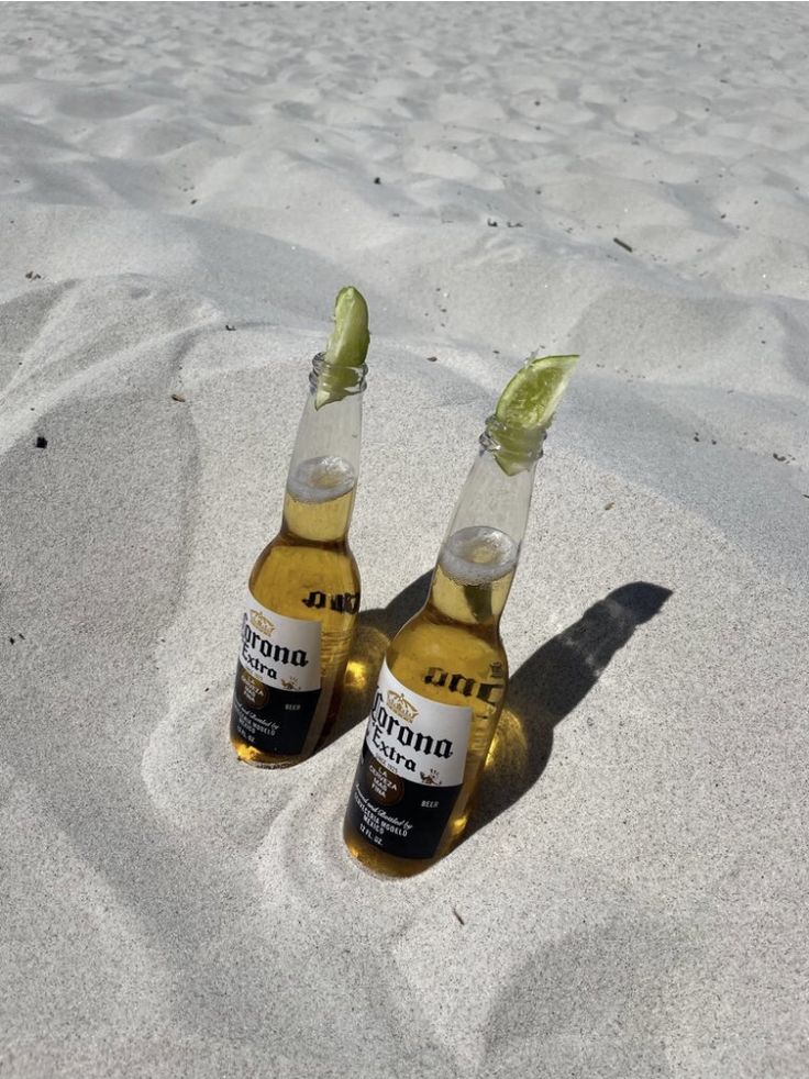 two bottles of beer sitting on top of a sandy beach next to the ocean with a lime wedge sticking out of one bottle