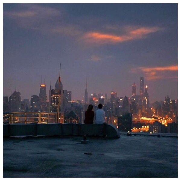 two people sitting on top of a roof with the city lights in the back ground