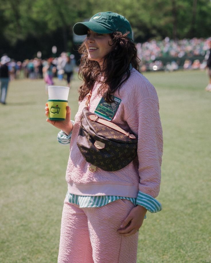 a woman in pink is holding a cup and wearing a fanny bag while standing on the grass