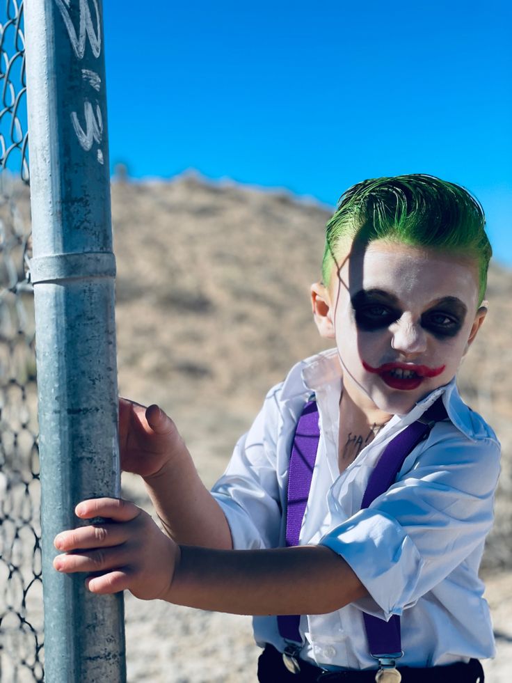 a young boy dressed as the joker leaning on a fence post with his face painted green