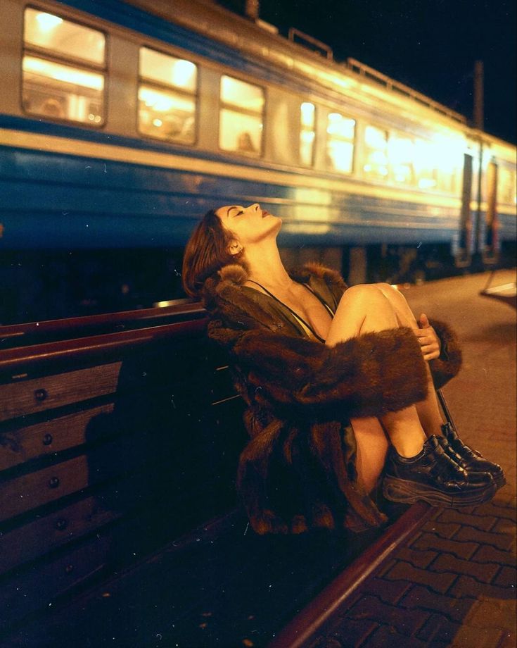 a woman sitting on a bench in front of a train at night with her eyes closed