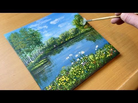 someone is painting a landscape with water and flowers on a wooden table, while holding a paintbrush