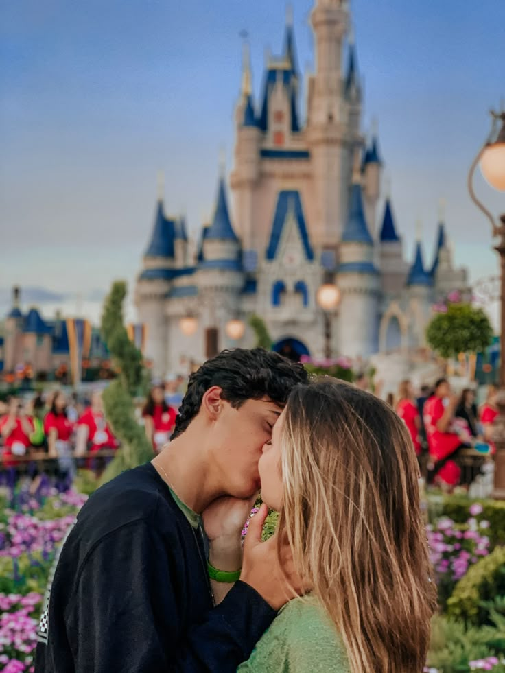 a young man and woman kissing in front of a castle at disney world with people standing around