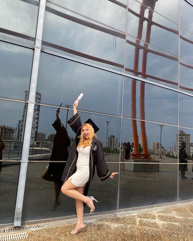 a woman is posing in front of a glass building with her graduation cap and gown on