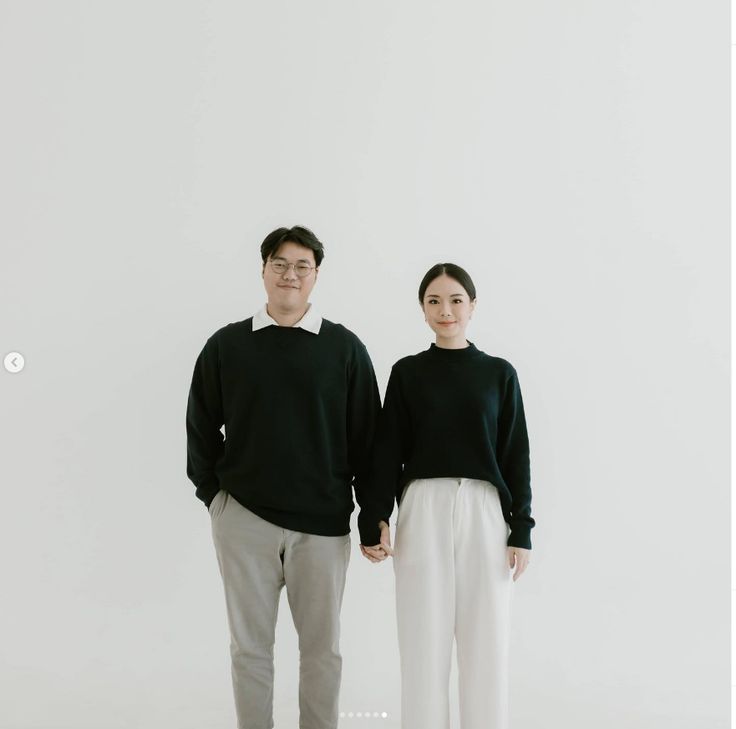 a man and woman standing next to each other in front of a white wall wearing black sweaters