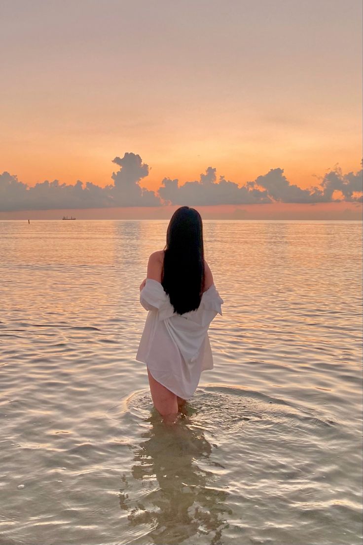 a woman standing in the water at sunset with her back to the camera, looking out into the distance