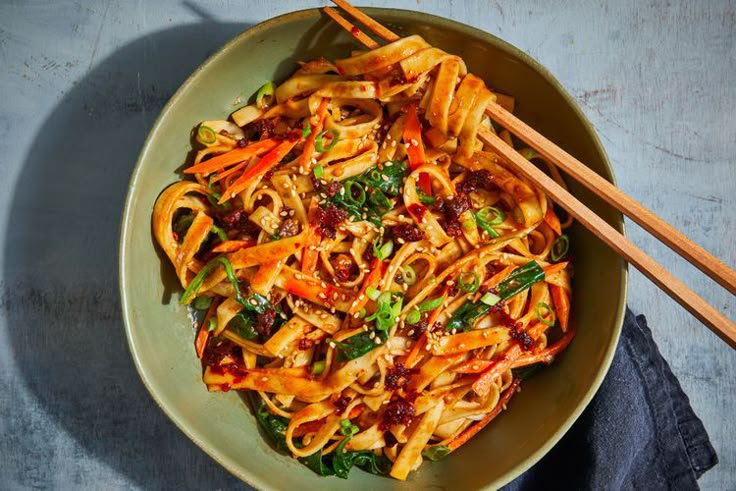 a bowl filled with noodles and vegetables next to chopsticks on a blue surface