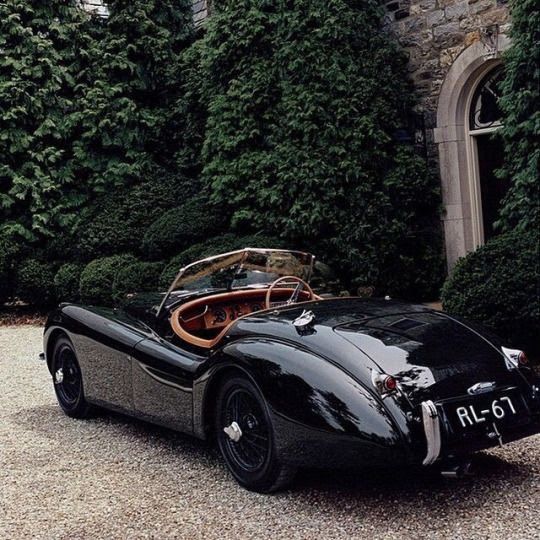 an old fashioned black sports car parked in front of a building with ivy covered walls