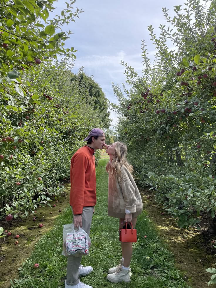 a man and woman standing in an apple orchard with one kissing the other's cheek