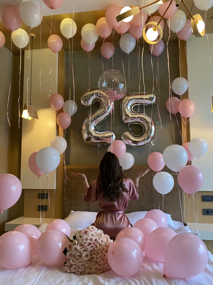 a woman sitting on top of a bed surrounded by pink and white balloons with the number twenty five