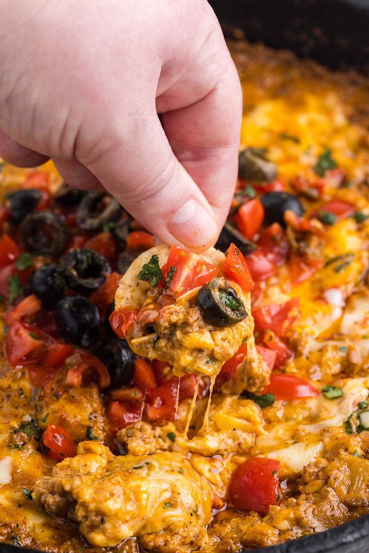 a hand is dipping some food into a skillet filled with cheese, tomatoes and black olives