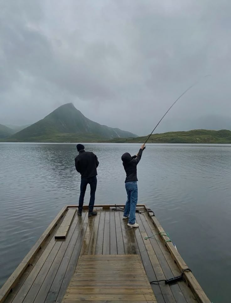 two people standing on a dock holding onto a fishing rod with mountains in the background