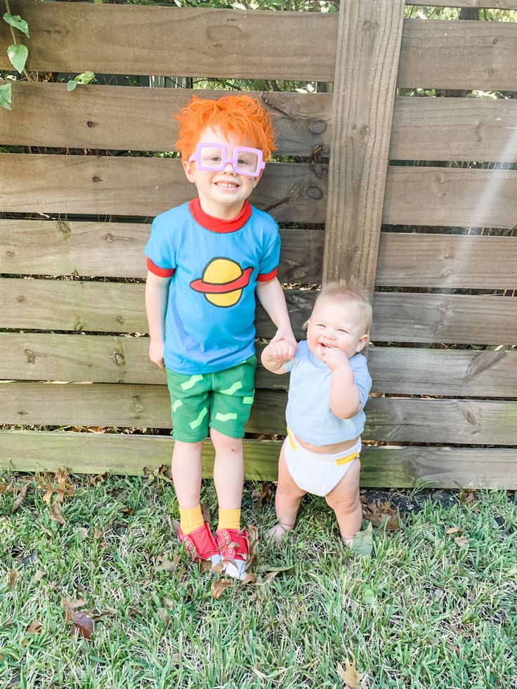 two young children standing next to each other in front of a wooden fence with grass