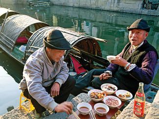 two men sitting on the edge of a dock eating food and drinking water from bowls