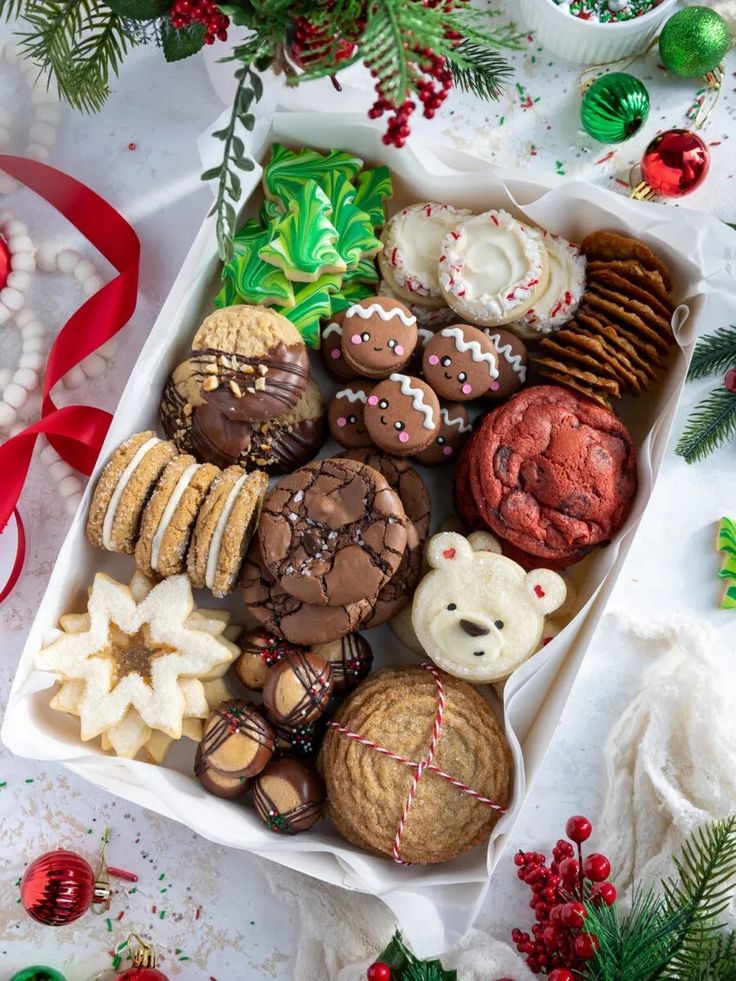 an assortment of christmas cookies in a white box with red ribbon and decorations around it