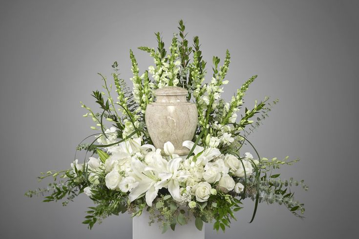 a vase filled with white flowers and greenery on top of a table next to a gray wall