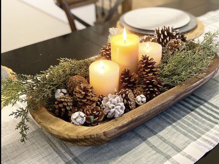 candles are lit in a wooden bowl filled with pine cones and greenery on a dining room table