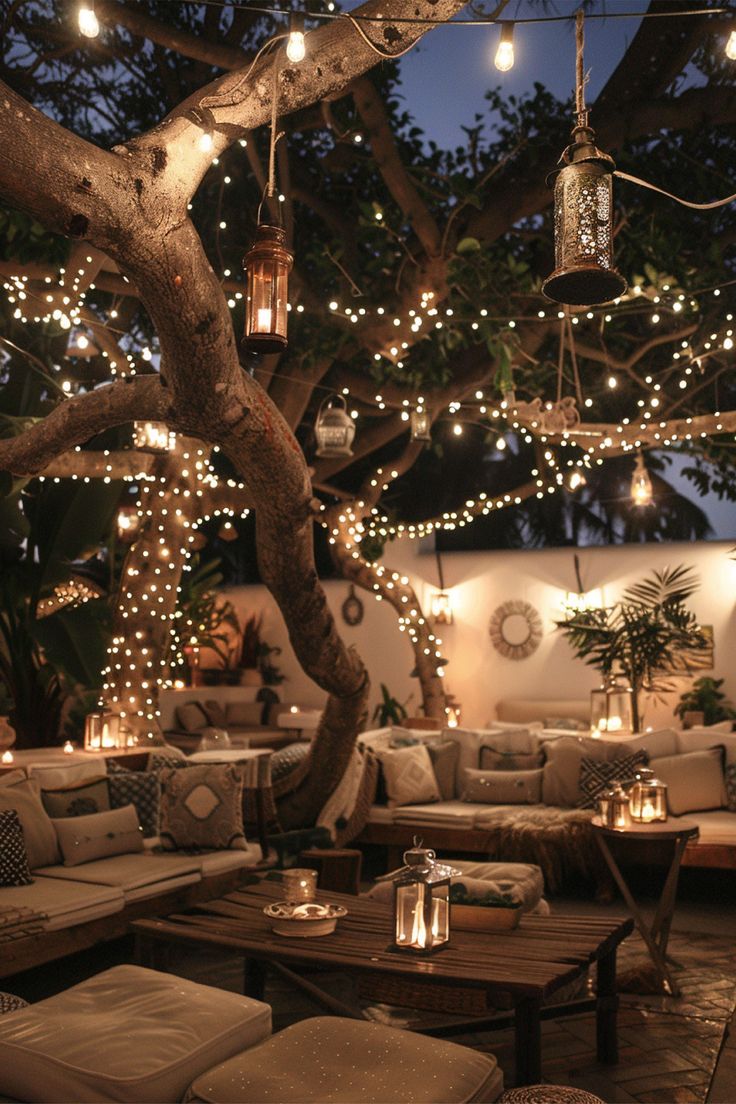 an outdoor seating area is lit up with string lights and hanging lanterns, which are hung from the branches of a tree