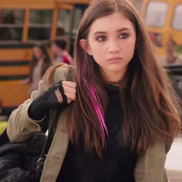 a young woman with long hair walking in front of a school bus and looking at the camera