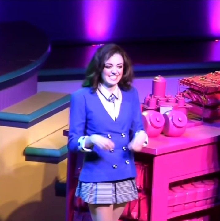 a woman standing in front of a pink table holding a nintendo wii game controller while wearing a blue blazer and skirt