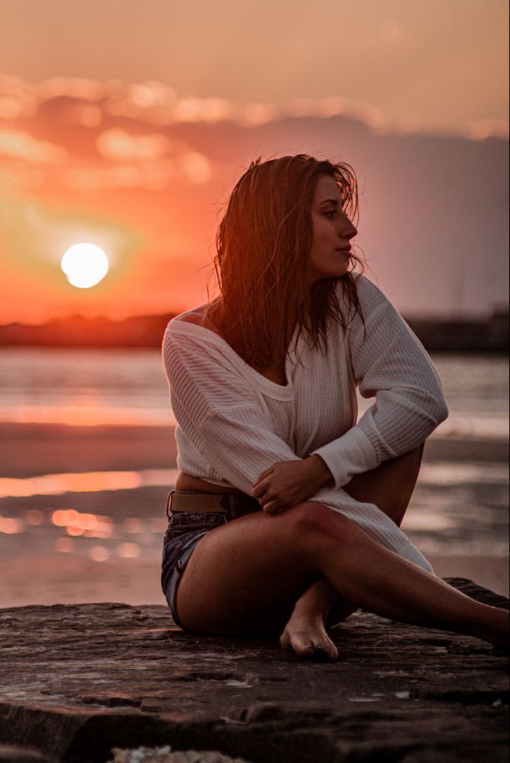 a woman sitting on top of a rock next to the ocean at sunset with her arms crossed