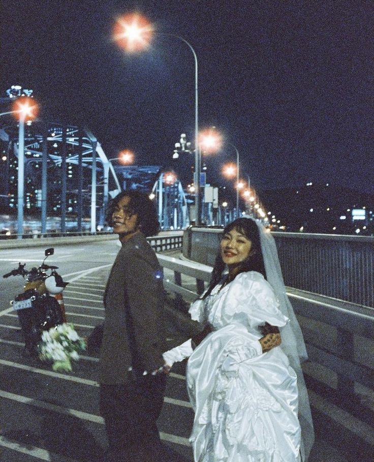 a bride and groom are standing on the bridge at night with their motorcycle behind them