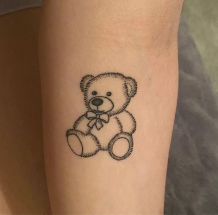 a small tattoo of a teddy bear on the right arm and lower leg, with a bow around it's neck