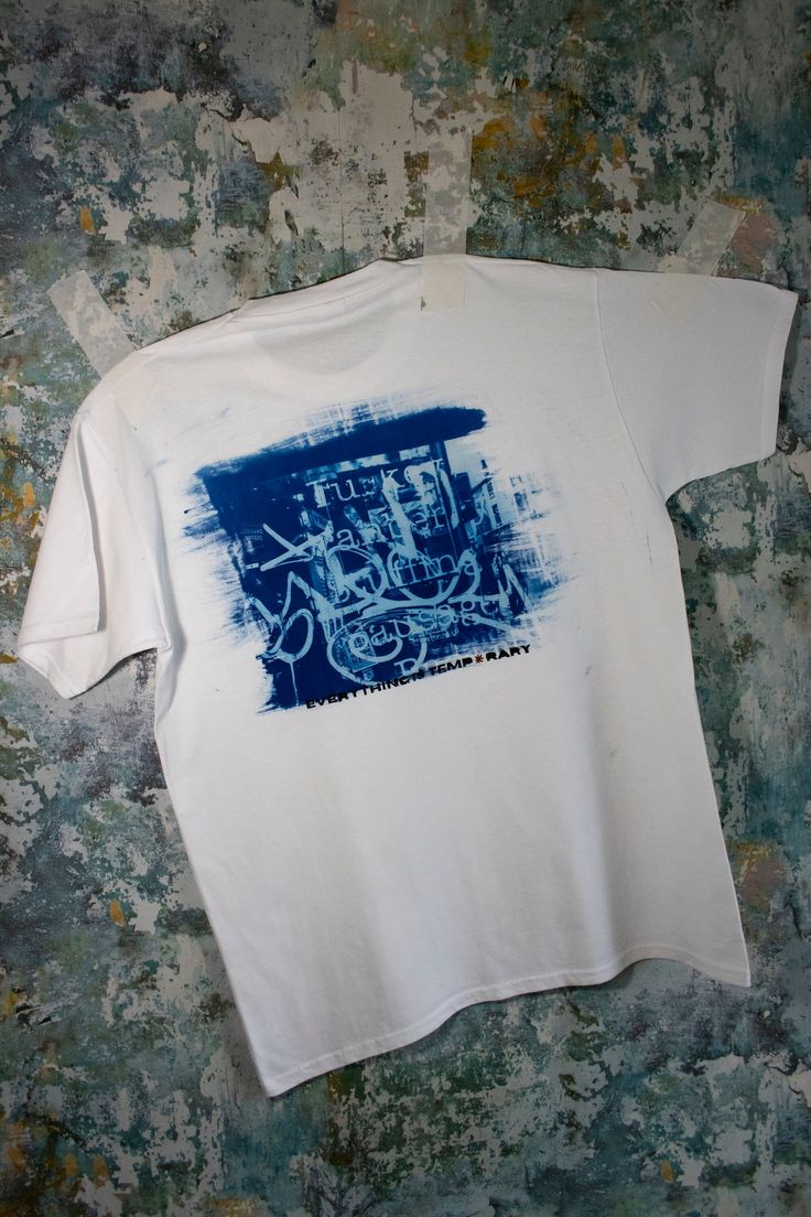 a white t - shirt with blue graffiti on the front and back, sitting against a wall