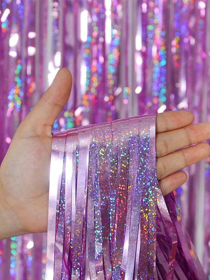 a hand is holding some purple streamers in front of a pink background with multicolored sequins