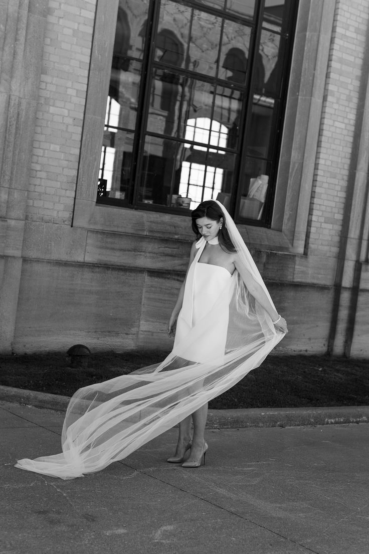 black and white photograph of woman in wedding dress with veil over head, standing on sidewalk