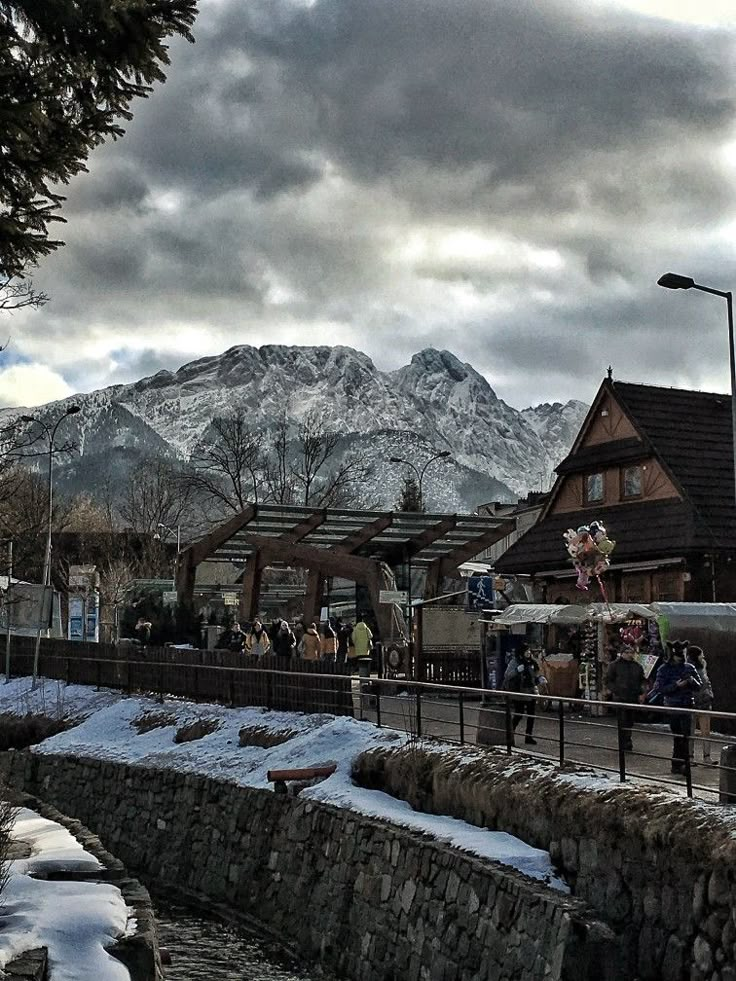 people are walking on the sidewalk in front of a mountain town with snow covered mountains behind them