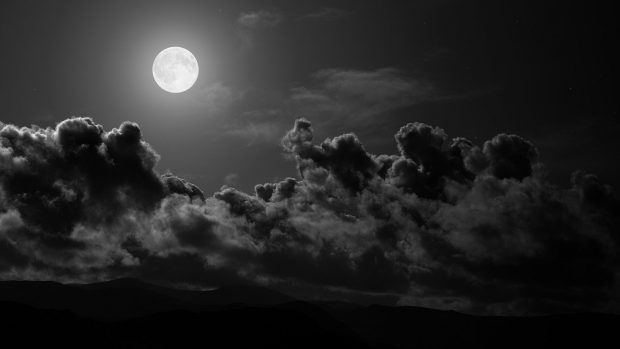 black and white photograph of clouds in the night sky with moon behind them on a cloudy day