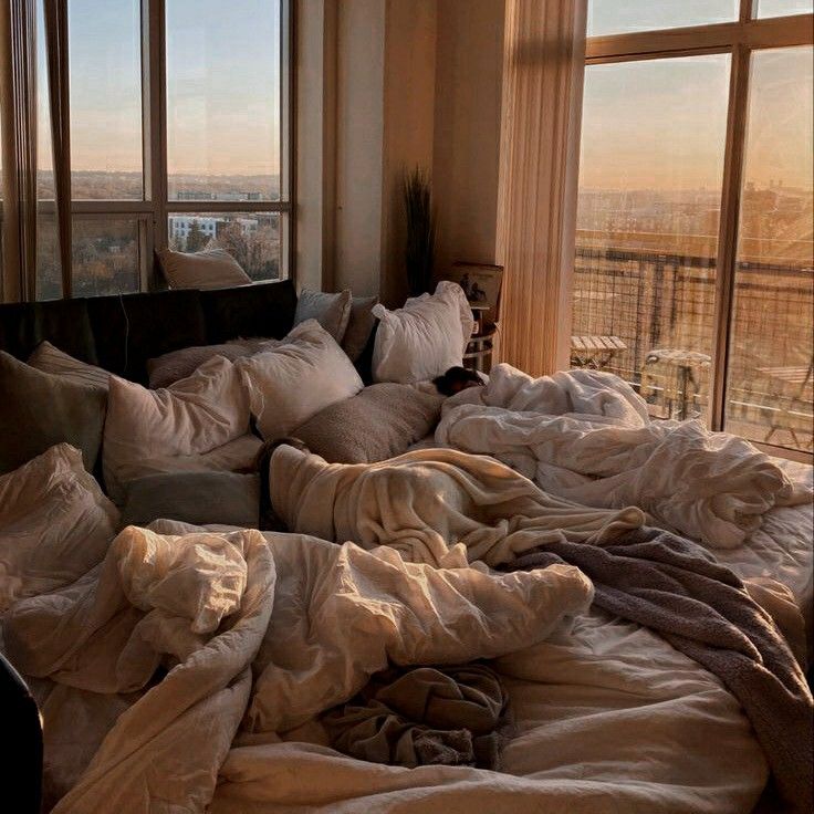 an unmade bed with many pillows and blankets on it in front of large windows