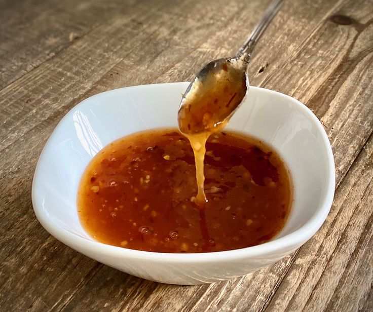 a spoon full of sauce being poured into a white bowl on top of a wooden table