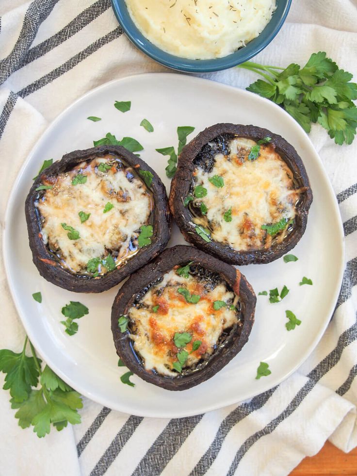 three stuffed mushrooms on a white plate next to a bowl of mashed potatoes and parsley