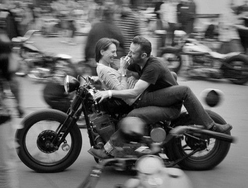 a man and woman riding on the back of a motorcycle down a street with motorcycles in the background