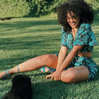 a woman sitting on the grass next to a black cat and smiling at the camera